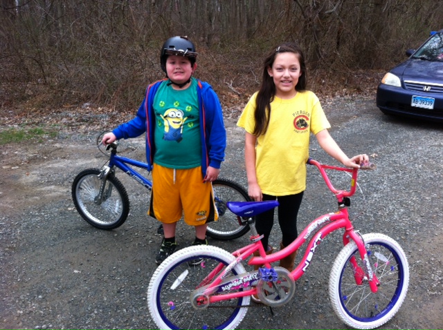 Two kids smiling with their bicycles outdoors on a paved path.